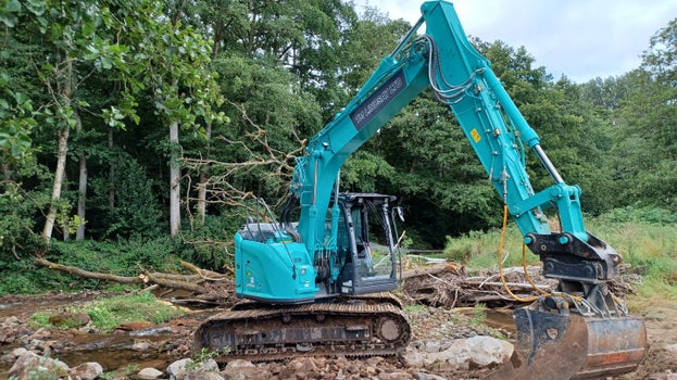 Turquoise digger excavating earth in a field on the edge of a river, with trees in the background